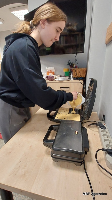 Osoba uczestnicząca w&nbsp;spotkaniu stoi przy stole kuchennym w&nbsp;bibliotece i&nbsp;przelewa porcję ciasta z&nbsp;miski do otwartej gofrownicy. W&nbsp;tle widoczne są szafki, akcesoria kuchenne oraz telewizor zawieszony na ścianie.