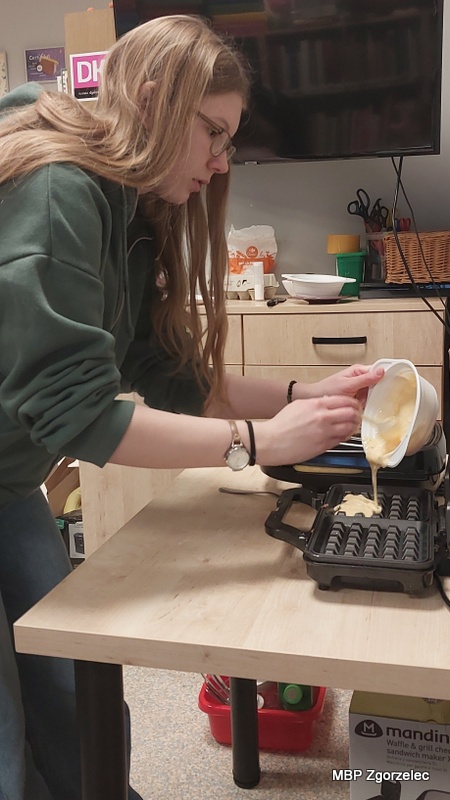 Osoba uczestnicząca w&nbsp;spotkaniu stoi przy stole kuchennym w&nbsp;bibliotece i&nbsp;przelewa porcję ciasta z&nbsp;miski do otwartej gofrownicy. W&nbsp;tle widoczne są szafki, akcesoria kuchenne oraz telewizor zawieszony na ścianie.