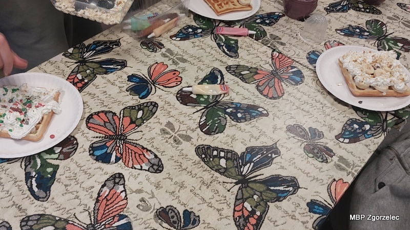 Na obrusie z&nbsp;motylami ustawiono papierowe talerzyki z&nbsp;świeżo upieczonymi goframi, udekorowanymi bitą śmietaną, posypkami i&nbsp;dodatkami. Obok widoczne są drobne akcesoria i&nbsp;składniki używane podczas kulinarnej części zajęć.
