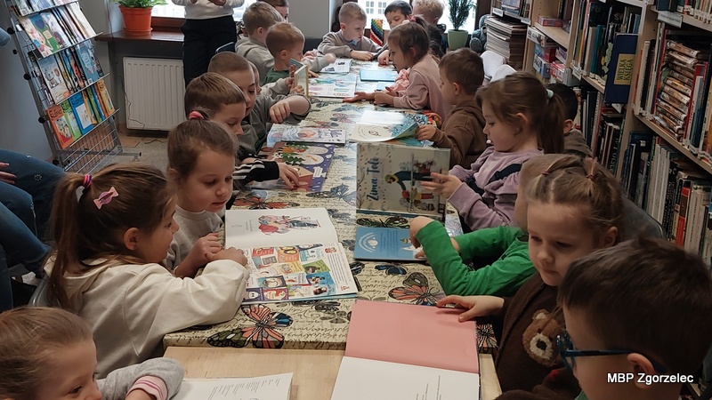 Grupa dzieci siedzi przy długim stole w bibliotece, przeglądając książki związane z tematyką zimy. Na stole leżą otwarte publikacje, a młodzi czytelnicy z zainteresowaniem oglądają ilustracje i rozmawiają o treści. W tle stoją regały z książkami, tworzące przytulną atmosferę bibliotecznych zajęć.