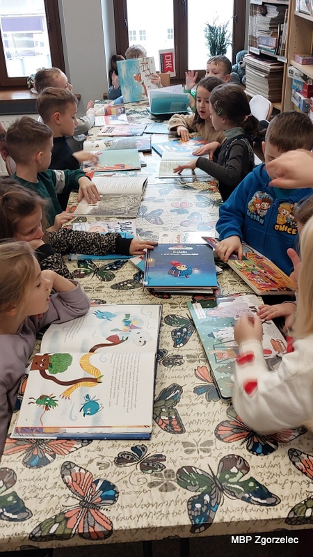 Grupa dzieci siedzi przy dużym stole w&nbsp;bibliotece, przeglądając kolorowe książki związane z&nbsp;tematyką zimy. Stół jest przykryty obrusem z&nbsp;motywem motyli, a&nbsp;przed każdym dzieckiem leży otwarta książka. W&nbsp;tle widać regały z&nbsp;książkami oraz jasne, duże okna wpuszczające naturalne światło, dzięki czemu atmosfera zajęć jest ciepła i&nbsp;sprzyjająca czytaniu. Dzieci uważnie oglądają ilustracje w&nbsp;książkach.