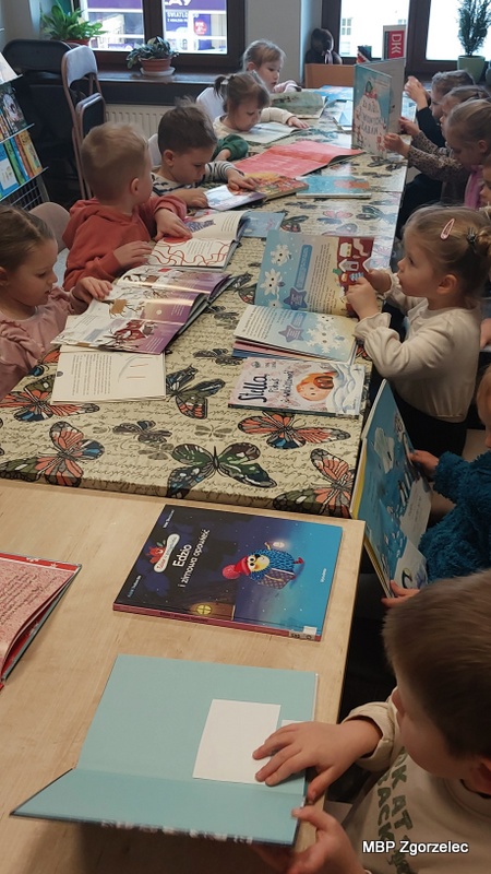 Grupa dzieci siedzi przy długim stole w&nbsp;bibliotece, przeglądając kolorowe książki dla najmłodszych. Na blacie leżą liczne otwarte publikacje z&nbsp;zimowymi ilustracjami i&nbsp;bajkami. W&nbsp;tle znajdują się regały oraz duże okna, a&nbsp;dzieci koncentrują się na oglądaniu ilustracji i&nbsp;stron książek jako część zajęć literackich.