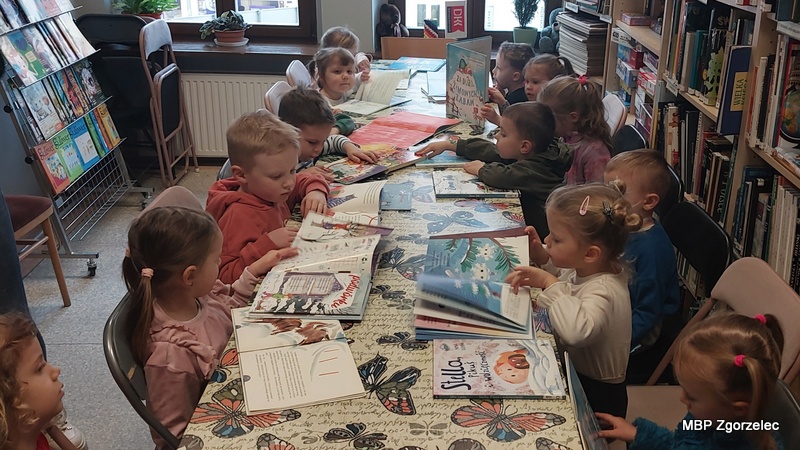 Dzieci siedzą przy długim stole w&nbsp;bibliotece, przeglądając otwarte książki z&nbsp;kolorowymi ilustracjami. Na stole rozłożone są różne publikacje dla najmłodszych, a&nbsp;uczestnicy zajęć wertują strony i&nbsp;oglądają rysunki. W&nbsp;otoczeniu widoczne są biblioteczne regały oraz stojak z&nbsp;czasopismami.