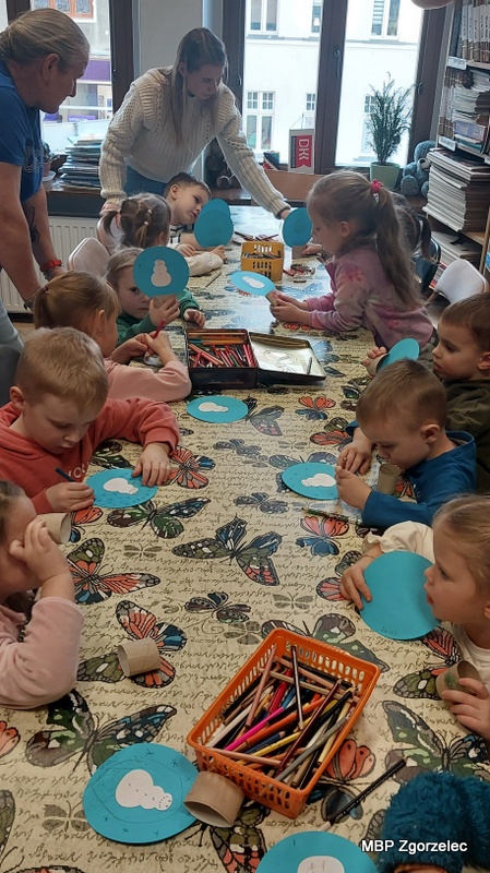 Dzieci siedzą przy dużym stole w&nbsp;bibliotece, trzymając w&nbsp;rękach niebieskie papierowe koła przygotowane do zimowych prac plastycznych. Na stole leżą kredki, materiały dekoracyjne oraz kleje. Dorośli stoją przy dzieciach i&nbsp;pomagają im rozpocząć tworzenie papierowych ozdób o&nbsp;tematyce zimowej.