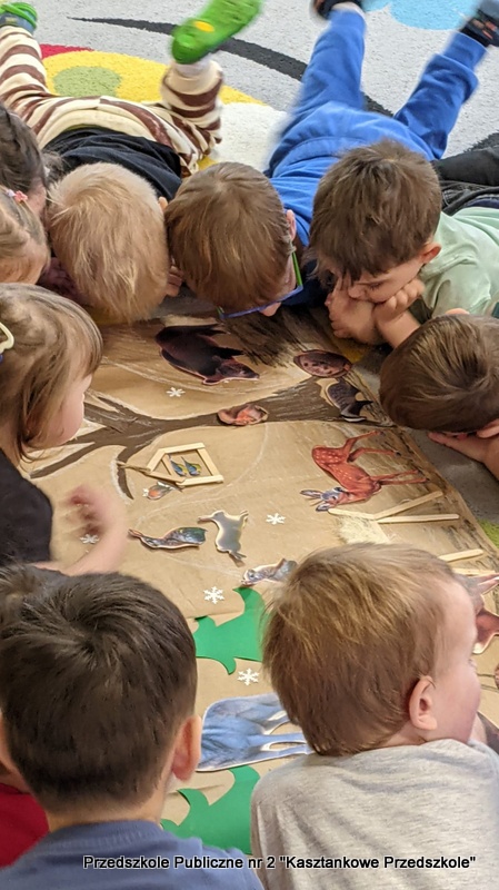 Grupa dzieci leży i&nbsp;siedzi wokół dużej, wspólnie wykonanej ilustracji (plakatu) rozłożonej na podłodze w&nbsp;przedszkolu. Ilustracja przedstawia zimową scenę z&nbsp;leśnymi zwierzętami, domkami i&nbsp;elementami krajobrazu. Dzieci pochylają się nad obrazkiem i&nbsp;wskazują różne szczegóły, tworząc wspólną aktywność poznawczą podczas zajęć.