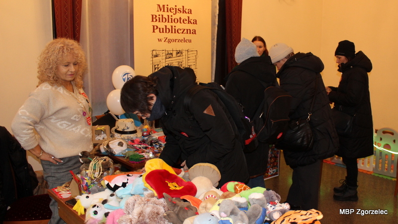 Stoisko MBP z&nbsp;pluszakami, drobnymi fantami i&nbsp;materiałami na loterię fantową. Odwiedzający przeglądają rzeczy wystawione na stole w&nbsp;ramach finału WOŚP.