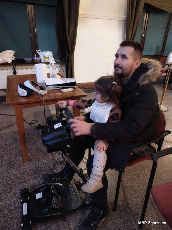 Rodzic z&nbsp;dzieckiem siedzą przy symulatorze jazdy wyposażonym w&nbsp;kierownicę i&nbsp;pedały. Dziecko trzyma kierownicę, korzystając z&nbsp;atrakcji MBP zorganizowanej w&nbsp;czasie finału WOŚP.