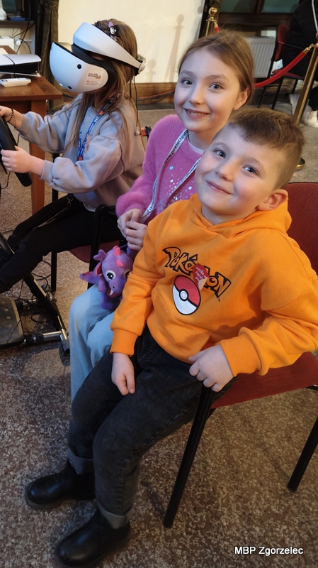 Grupa dzieci siedzi na krzesłach i&nbsp;korzysta z&nbsp;symulatora jazdy w&nbsp;goglach VR, obserwując wirtualny tor wyścigowy podczas atrakcji przygotowanych przez MBP.
