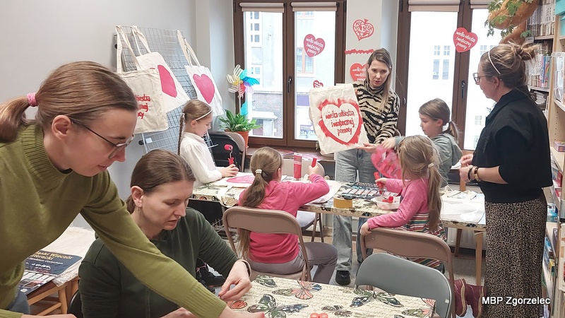 Grupa dzieci i&nbsp;prowadzących podczas warsztatów zdobienia ekologicznych toreb z&nbsp;motywem WOŚP w&nbsp;bibliotece. Na stole leżą farby, pędzle i&nbsp;gotowe torby.