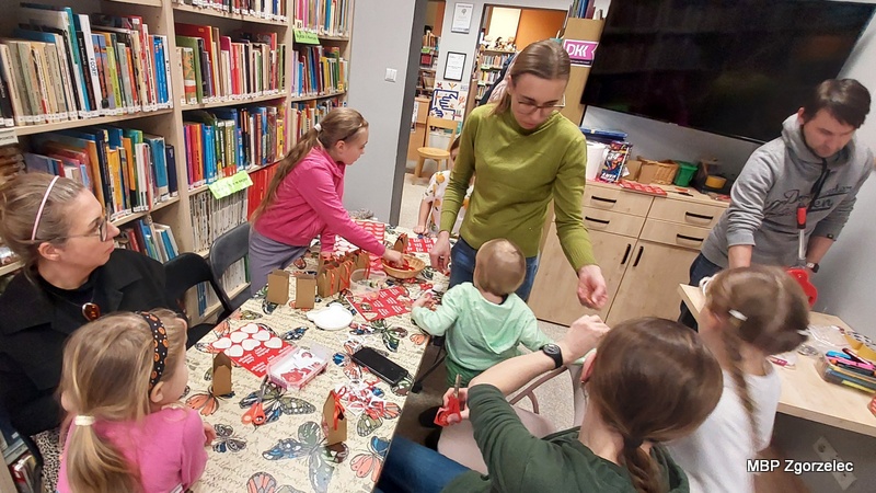 Uczestnicy warsztatów w&nbsp;bibliotece przygotowują przypinki WOŚP oraz małe upominki, pracując wspólnie przy dużym stole wśród półek z&nbsp;książkami.