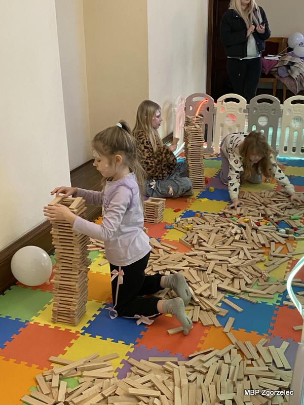 Dzieci bawią się dużą liczbą drewnianych klocków na kolorowej piankowej macie, budując wysokie wieże podczas atrakcji zorganizowanych przez MBP w&nbsp;ramach finału WOŚP.