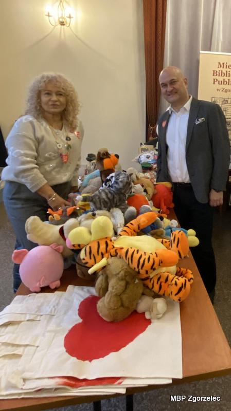 Pani Krystyna Radzięta - Dyrektor MBP w&nbsp;Zgorzelcu i&nbsp;Pan Rafał Gronicz - Burmistrz Miasta Zgorzelec stoją przy stole wypełnionym maskotkami i&nbsp;materiałami WOŚP, prezentując stoisko biblioteki w&nbsp;Miejskim Domu Kultury.