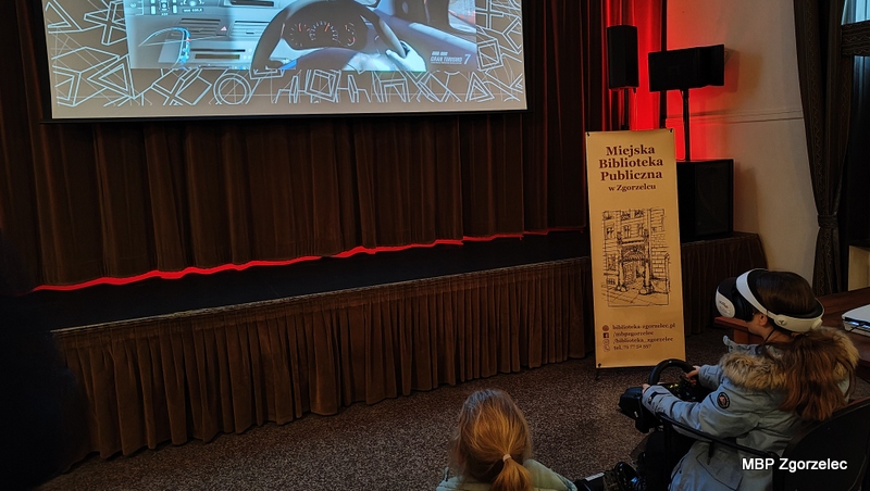 Uczestnicy w&nbsp;Miejskim Domu Kultury obserwują rozgrywkę z&nbsp;gry Gran Turismo 7 wyświetloną na dużym ekranie. Obok stoi roll‑up Miejskiej Biblioteki Publicznej, a&nbsp;młodzi uczestnicy próbują wirtualnej jazdy samochodem.