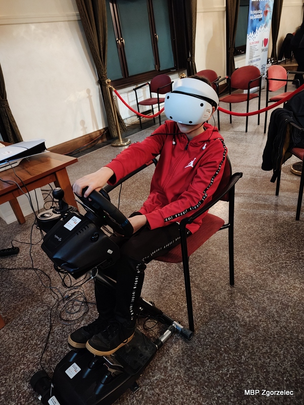 Młody chłopiec korzysta z&nbsp;symulatora jazdy VR — siedzi przed kierownicą i&nbsp;pedałami, mając na głowie gogle VR.