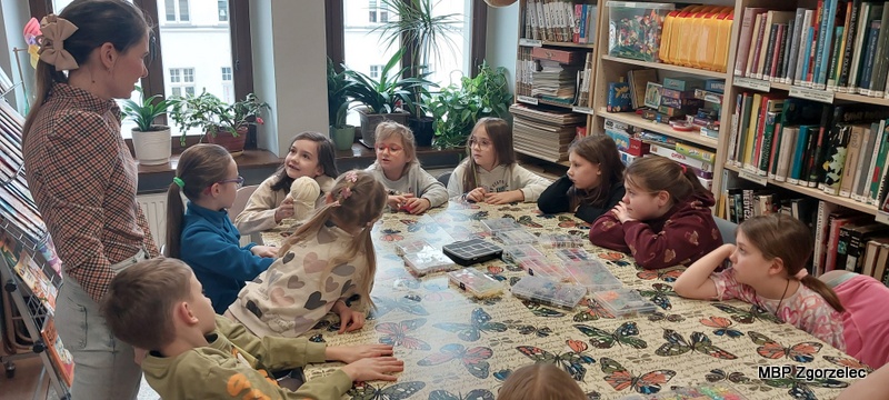 Przy dużym stole pokrytym obrusem z&nbsp;motylami siedzi grupa dzieci uczestniczących w&nbsp;zajęciach plastycznych w&nbsp;Miejskiej Bibliotece Publicznej w&nbsp;Zgorzelcu. Na blacie leżą pojemniki z&nbsp;elementami do tworzenia prac oraz narzędzia plastyczne. Obok dzieci stoi prowadząca zajęcia. W&nbsp;tle widoczne są regały z&nbsp;książkami, gry edukacyjne i&nbsp;okna z&nbsp;roślinami.