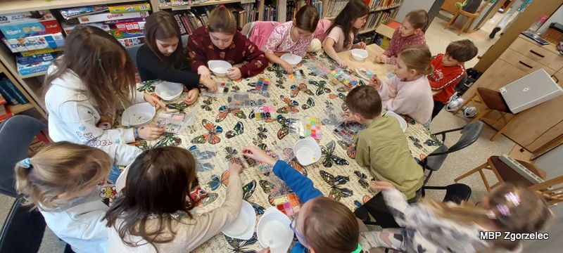 Dzieci siedzą wokół dużego stołu przykrytego obrusem z&nbsp;motylami i&nbsp;tworzą prace plastyczne z&nbsp;wykorzystaniem wielu drobnych elementów i&nbsp;miseczek ustawionych przed nimi. Każde dziecko pracuje nad własnym projektem. W&nbsp;tle widoczne są regały z&nbsp;książkami oraz część działu gier planszowych.