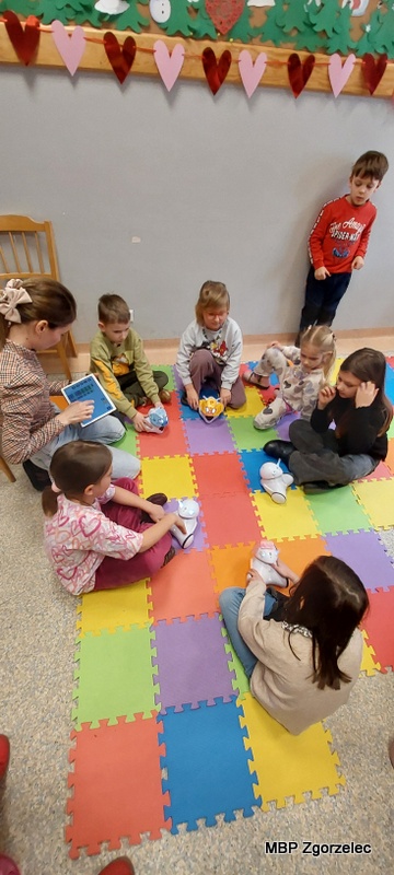 Dzieci siedzą w&nbsp;kole na kolorowych matach piankowych, uczestnicząc w&nbsp;zajęciach wprowadzających do programowania i&nbsp;logicznego myślenia. Używają interaktywnych robotów oraz tableta, którą trzyma prowadząca. W&nbsp;tle na ścianie zawieszona jest dekoracja z&nbsp;serduszkami, a&nbsp;obok stoi chłopiec obserwujący zajęcia.