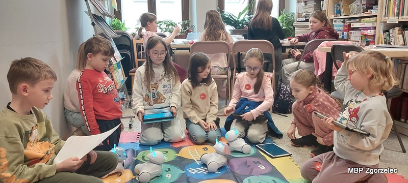 Na kolorowej macie edukacyjnej siedzi grupa dzieci, które pracują z&nbsp;małymi robotami wyposażonymi w&nbsp;świecące elementy. Niektóre dzieci korzystają z&nbsp;tabletów, inne trzymają kartki z&nbsp;zadaniami. W&nbsp;tle znajduje się długi stół, przy którym pracuje kolejna grupa uczestników, oraz regały z&nbsp;książkami i&nbsp;grami.