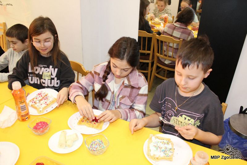Przy żółtym stole dzieci dekorują gofry różnymi słodkimi dodatkami. Na stole znajdują się talerzyki, miseczki z&nbsp;posypkami, bita śmietana, owoce i&nbsp;napoje. Każde dziecko pracuje nad własnym deserem w&nbsp;przyjaznej przestrzeni biblioteki.