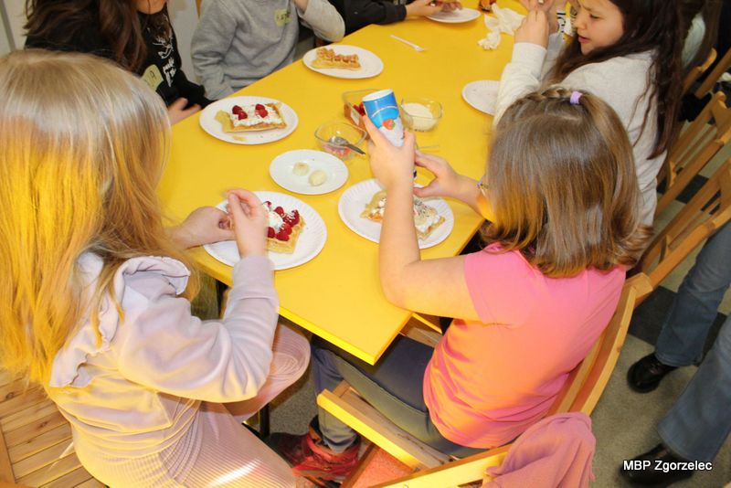 Przy żółtym stole dzieci tworzą słodkie przekąski, nakładając na gofry owoce, kremy i&nbsp;kolorowe posypki. Na blacie leżą talerzyki z&nbsp;przygotowanymi waflami oraz małe miski z&nbsp;dodatkami do dekorowania. Zajęcia mają charakter wspólnej zabawy kulinarnej w&nbsp;bibliotece.