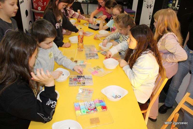 Grupa dzieci pracuje przy długim żółtym stole, wybierając kolorowe koraliki, zawieszki i&nbsp;elementy do bransoletek oraz naszyjników. Na stole leżą przegródkowe pojemniki z&nbsp;materiałami plastycznymi oraz miseczki przygotowane do pracy. Warsztaty odbywają się w&nbsp;przestrzeni bibliotecznej z&nbsp;elementami dekoracyjnymi wokół.