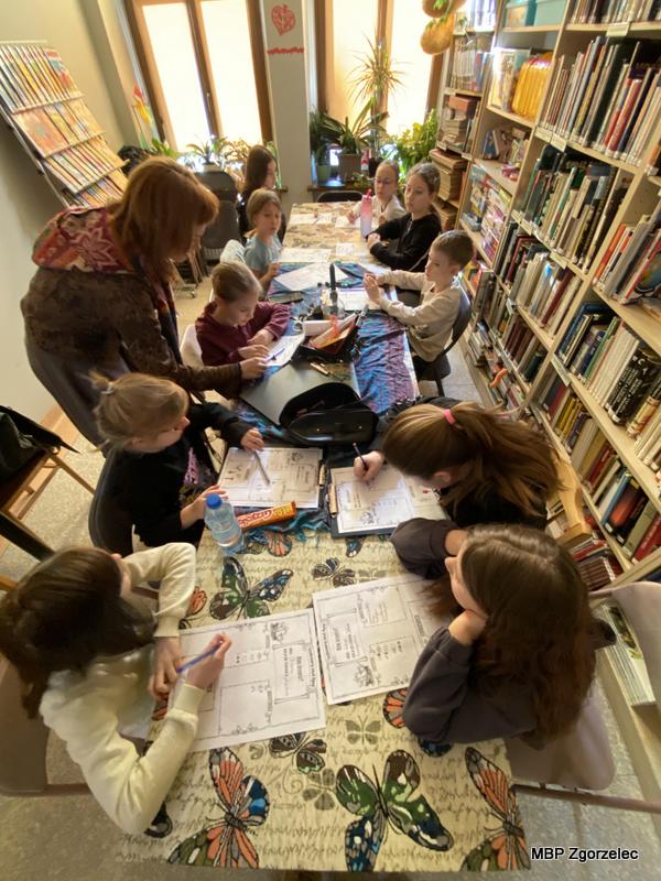 Przy długim stole pokrytym obrusem w&nbsp;motyle siedzi grupa dzieci, które wypełniają karty pracy z&nbsp;zadaniami edukacyjnymi. Prowadząca stoi obok i&nbsp;pomaga jednemu z&nbsp;uczestników. W&nbsp;tle widać regały pełne książek oraz duże okna, przez które wpada światło.