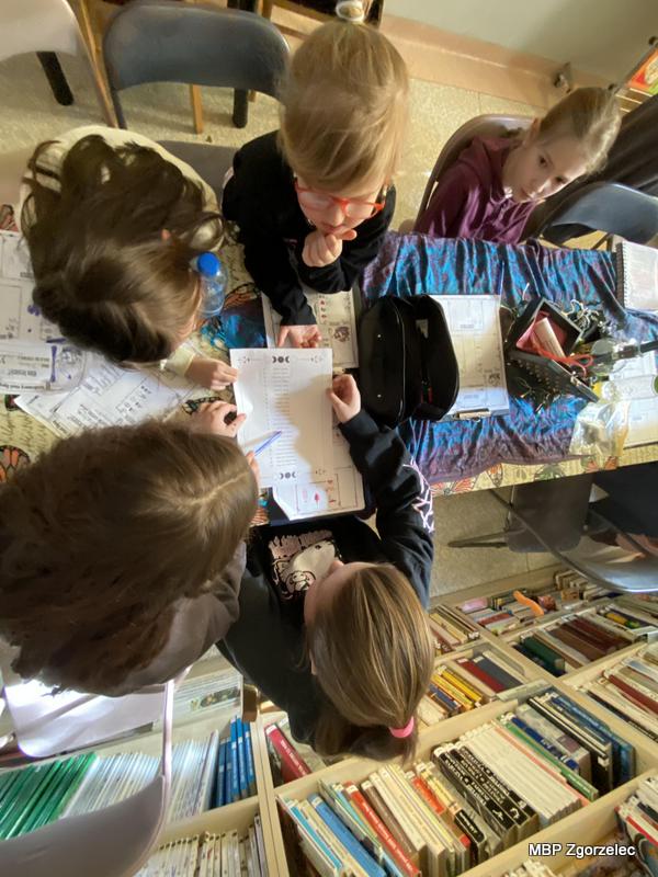 Ujęcie z&nbsp;góry przedstawia grupę dzieci pochylonych nad kartami pracy. Dzieci współpracują, rysują i&nbsp;zapisują odpowiedzi. Na stole leżą ołówki, długopisy, kolorowe drobiazgi, tornistry oraz materiały wykorzystywane podczas zajęć. Wokół stołu widoczne są regały z&nbsp;książkami.