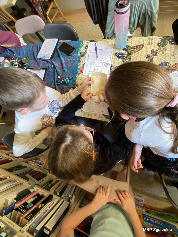 Kilka dzieci stoi i&nbsp;siedzi przy stole, analizując kartkę z&nbsp;treścią zadania lub wskazówkami. Na stole znajdują się karty pracy, przybory do pisania, butelki z&nbsp;wodą i&nbsp;notesy. W&nbsp;tle widać niskie regały z&nbsp;książkami i&nbsp;materiały przygotowane na zajęcia edukacyjne.
