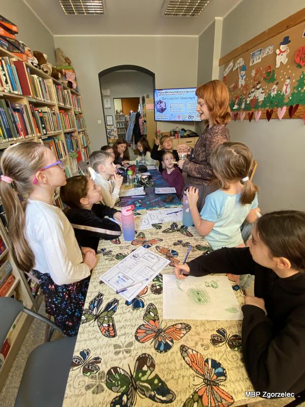 Dzieci siedzą przy stole z&nbsp;kartami pracy i&nbsp;przyborami do pisania, podczas gdy prowadząca omawia elementy gry fabuarnej. Na ścianie z&nbsp;tyłu widnieje dekoracja z&nbsp;kolorowymi sercami oraz telewizor. W&nbsp;tle stoją regały z&nbsp;książkami działu dziecięcego oraz plansze tematyczne.