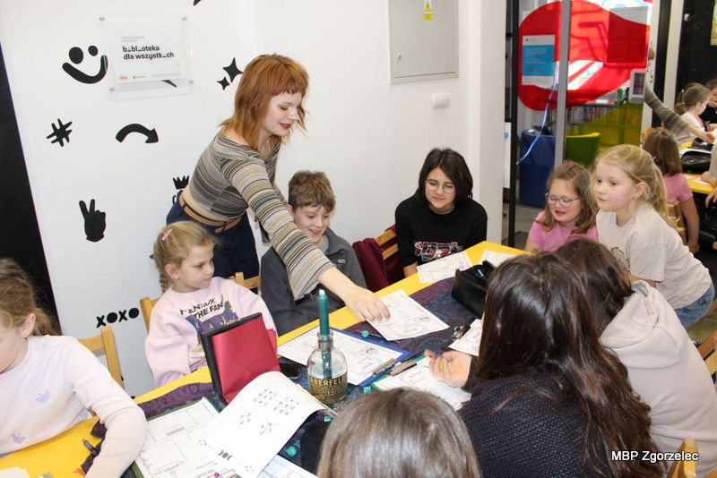 Dzieci siedzą przy stole, pracując na kartach z&nbsp;ćwiczeniami, a&nbsp;prowadząca pochyla się nad jednym z&nbsp;uczestników, przekazując mu materiał lub wskazówkę. Na stole znajdują się zeszyty, notatki, długopisy oraz przybory do pracy. W&nbsp;tle widać dekoracje ścienne i&nbsp;wejście do sąsiedniej części biblioteki.