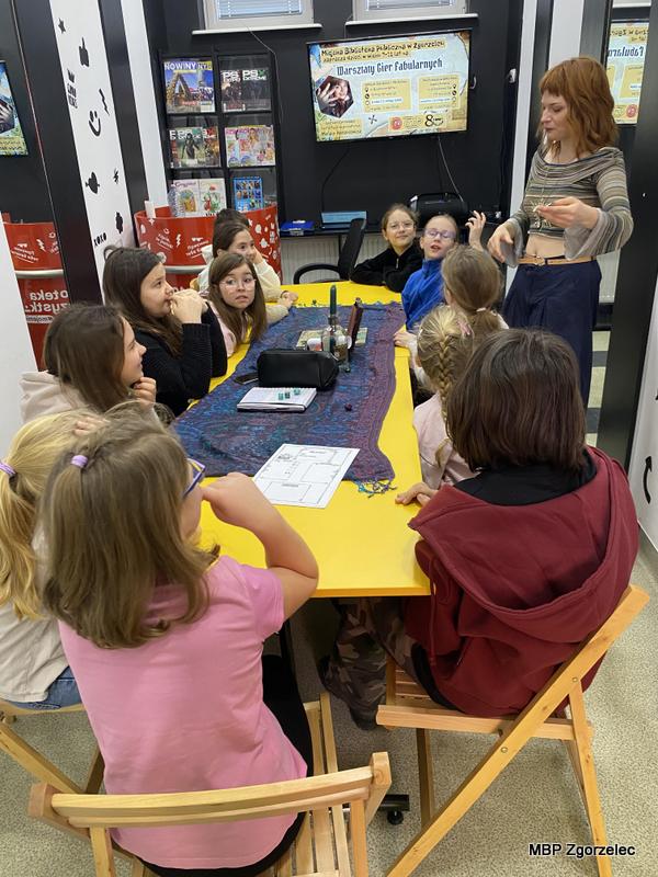 W sali bibliotecznej dzieci siedzą przy długim, żółtym stole, na którym leżą karty pracy, ołówki i&nbsp;materiały dydaktyczne. Przed stołem stoi prowadząca, która gestykuluje i&nbsp;omawia grę fabularną. W&nbsp;tle widoczne są plakaty, telewizor oraz regał z&nbsp;czasopismami.