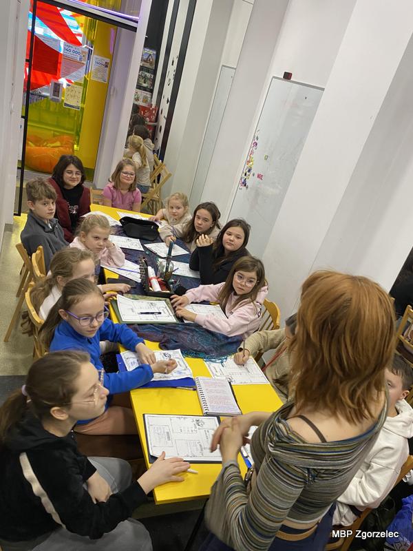 W przestrzeni bibliotecznej dzieci siedzą przy dwóch połączonych stołach, na których leżą karty pracy, piórniki i&nbsp;notatki. Prowadząca stoi przy stole i&nbsp;omawia grę fabularną. W&nbsp;tle widoczne są kolorowe elementy dekoracyjne i&nbsp;wejście do innej części Mediateki.