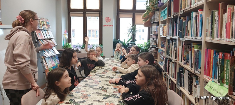 Przy długim stole w&nbsp;dziale dziecięcym biblioteki siedzi grupa dzieci uczestniczących w&nbsp;zajęciach edukacyjnych. Kilkoro z&nbsp;nich podnosi rękę, odpowiadając na pytanie prowadzącej stojącej z&nbsp;boku stołu. W&nbsp;tle widać regały z&nbsp;książkami, stojak z&nbsp;czasopismami i&nbsp;duże okna oświetlające przestrzeń.