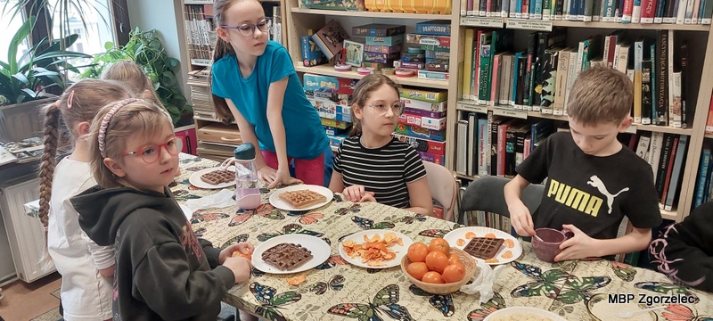 Dzieci siedzą przy stole w&nbsp;dziale dziecięcym biblioteki, gdzie przygotowują gofry, dodając owoce, posypki i&nbsp;inne składniki. Na środku stołu stoją miseczki z&nbsp;owocami oraz talerze z&nbsp;waflami. W&nbsp;tle znajdują się regały z&nbsp;książkami i&nbsp;gry edukacyjne ustawione na półkach.
