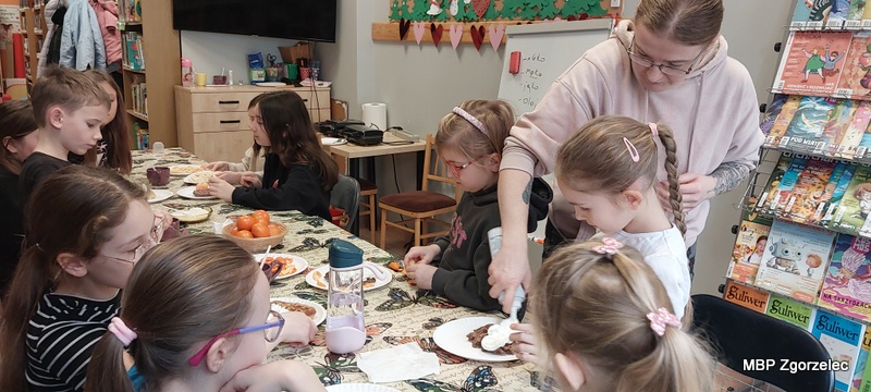Przy stole przykrytym kolorowym obrusem dzieci przygotowują gofry. Prowadząca trzyma pojemnik z&nbsp;bitą śmietaną i&nbsp;pomaga jednemu z&nbsp;dzieci wykonać deser. Na stole znajdują się talerzyki z&nbsp;goframi, owoce, dekoracje spożywcze oraz napoje. W&nbsp;tle widać dekoracje ścienne oraz regał z&nbsp;książkami i&nbsp;czasopismami.