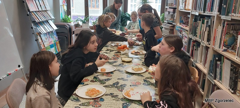 Dzieci siedzą przy stole i&nbsp;jedzą przygotowane wcześniej gofry oraz owoce rozłożone na talerzykach. Na stole znajduje się kilka naczyń z&nbsp;dodatkami oraz owocami. W&nbsp;tle widoczne są regały z&nbsp;książkami i&nbsp;okna wychodzące na ulicę.