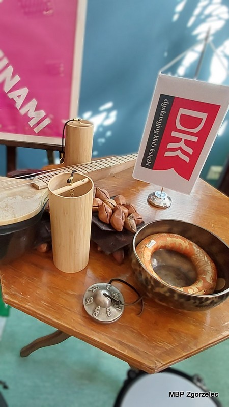Na stole leżą różnorodne instrumenty muzyczne, w&nbsp;tym misa dźwiękowa, instrument strunowy, bambusowy deszczowiec, małe talerzyki oraz inne przedmioty wykorzystywane podczas muzycznych eksperymentów. Obok znajduje się mała flaga Dyskusyjnego Klubu Książki.