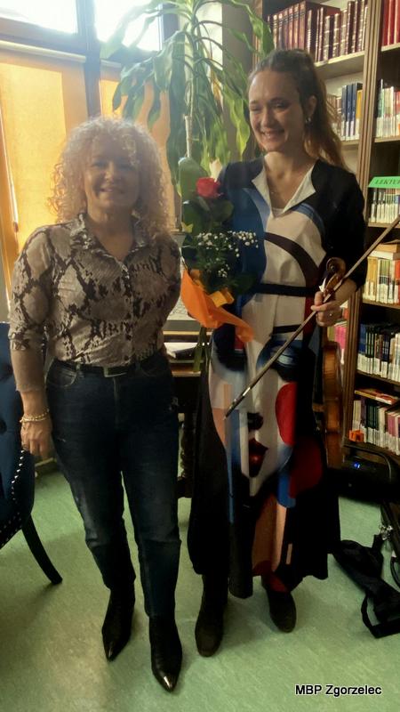 Dyrekcja MBP Zgorzelec - Krystyna Radzięta i&nbsp;Pani Justyna Miguła stoi w&nbsp;Czytelni trzymając skrzypce i&nbsp;bukiet kwiatów po zakończonych warsztatach. W&nbsp;tle znajdują się regały z&nbsp;książkami.