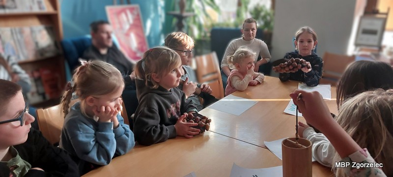 Dzieci siedzą przy stole i&nbsp;trzymają w&nbsp;dłoniach proste instrumenty etniczne, biorąc udział w&nbsp;muzycznych eksperymentach. Na blacie stołu leżą kartki i&nbsp;misa dźwiękowa używana podczas zajęć.