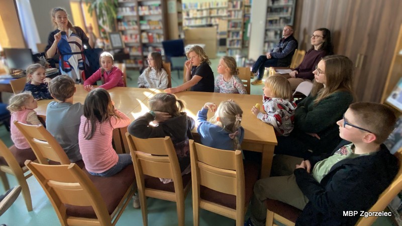 Grupa dzieci i opiekunów siedzi wokół dużego stołu w bibliotece podczas warsztatów „Literatura w dźwiękach”. Prowadząca stoi obok stołu i opowiada o muzyce oraz nadchodzących zadaniach. W tle widoczne są regały z książkami i stanowiska komputerowe.