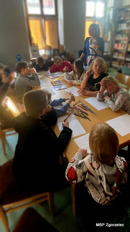 Dzieci siedzą przy stole z&nbsp;rozłożonymi kartkami i&nbsp;kredkami, a&nbsp;prowadząca stoi obok i&nbsp;wyjaśnia kolejne ćwiczenie związane z&nbsp;tworzeniem rysunkowych interpretacji muzyki. Światło wpada przez duże okna biblioteki.