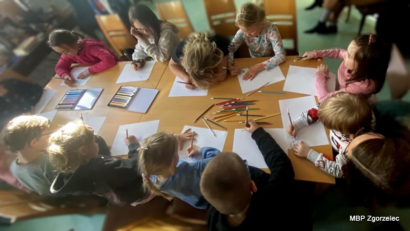 Grupa dzieci pochyla się nad stołem, rysując ołówkami i&nbsp;kredkami w&nbsp;ramach ćwiczeń łączących muzykę z&nbsp;wyobraźnią. Na stole leżą kartki i&nbsp;zestawy kredek, a&nbsp;uczestnicy intensywnie pracują nad ilustracjami inspirowanymi dźwiękiem.