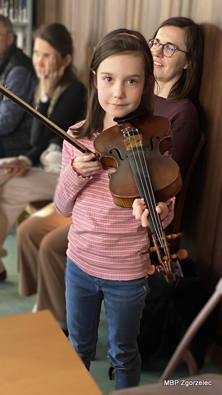 Dziewczynka stoi w&nbsp;bibliotece, trzymając w&nbsp;dłoniach skrzypce i&nbsp;smyczek podczas prezentacji instrumentów. W&nbsp;tle siedzą opiekunowie i&nbsp;pozostali uczestnicy warsztatów.