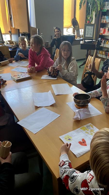 Dzieci siedzą wokół stołu, na którym leżą kartki i&nbsp;misa dźwiękowa. Jedno z&nbsp;dzieci pokazuje własny rysunek powstały w&nbsp;ramach ćwiczeń z&nbsp;twórczej interpretacji muzyki. Atmosfera jest skupiona i&nbsp;twórcza.