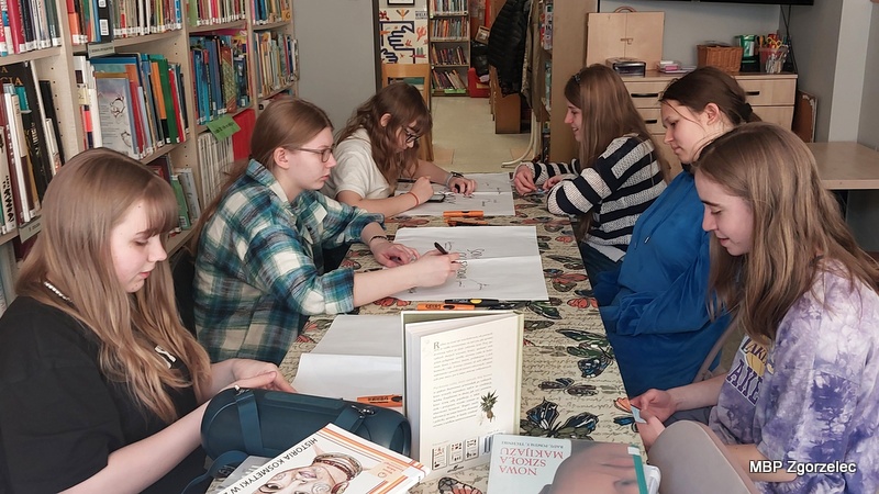 Grupa młodzieży z Młodzieżowego Klubu Literackiego „Książkowi Wędrowcy” siedzi przy dużym stole w czytelni biblioteki i przygotowuje mapę myśli o zastosowaniu: soli epsom, sody oczyszczonej i kwasku cytrynowego związane z warsztatami. Wokół znajdują się książki, w tym publikacje o naturalnych kosmetykach i ziołach. 