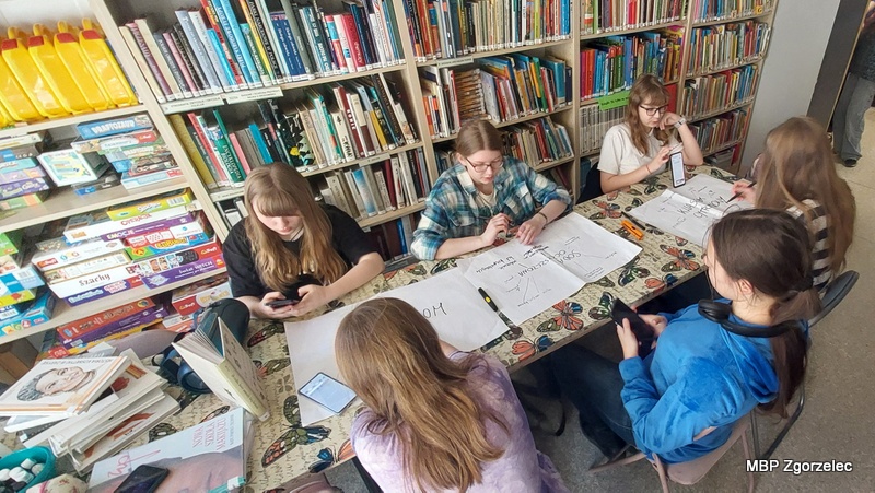 Grupa młodych klubowiczów siedzi przy stole w&nbsp;otoczeniu bibliotecznych regałów, tworząc  mapę myśli o&nbsp;zastosowaniu: soli epsom, sody oczyszczonej i&nbsp;kwasku cytrynowego związane z&nbsp;warsztatami. Widać telefony, arkusze papieru i&nbsp;książki rozłożone na stole.