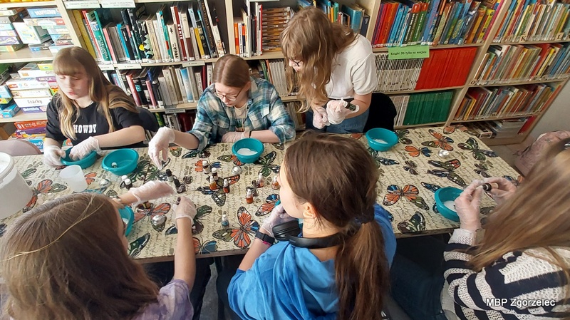 Uczestnicy warsztatów stoją i&nbsp;siedzą wokół stołu w&nbsp;Oddziale dla Dzieci i&nbsp;Młodzieży, wybierając aromaty oraz barwniki do tworzenia kul kąpielowych. Na stole znajdują się miski, rękawiczki, olejki eteryczne i&nbsp;podstawowe składniki kosmetyczne. Młodzież eksperymentuje z&nbsp;zapachami, korzystając jednocześnie z&nbsp;książek o&nbsp;kosmetyce dostępnych w&nbsp;zbiorach biblioteki jako inspiracji.