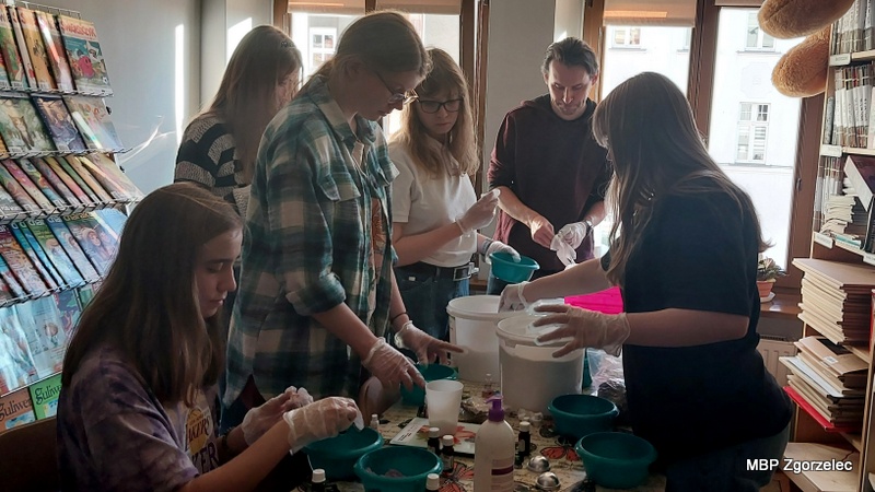 Grupa młodzieży pracuje zespołowo, mieszając składniki potrzebne do wykonania kul kąpielowych. Uczestnicy korzystają z&nbsp;przygotowanych materiałów i&nbsp;narzędzi, a&nbsp;na stole widać olejki aromatyczne, miski, miarki i&nbsp;butelki. Spotkanie ma twórczy charakter, a&nbsp;rozmowy nawiązują do obchodzonego Światowego Dnia Pisarzy i&nbsp;książek o&nbsp;kosmetykach znajdujących się w&nbsp;bibliotece.