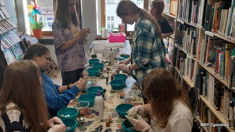 Uczestnicy warsztatów pracują nad formowaniem musujących kul kąpielowych. Każda osoba ma przed sobą miseczkę ze składnikami i&nbsp;tworzy własne kosmetyki, dobierając kolory i&nbsp;zapachy. Na stole znajdują się liczne olejki, dodatki i&nbsp;buteleczki, a&nbsp;atmosfera sprzyja eksperymentowaniu i&nbsp;rozmowom m.in. o&nbsp;książkach dotyczących naturalnej pielęgnacji.