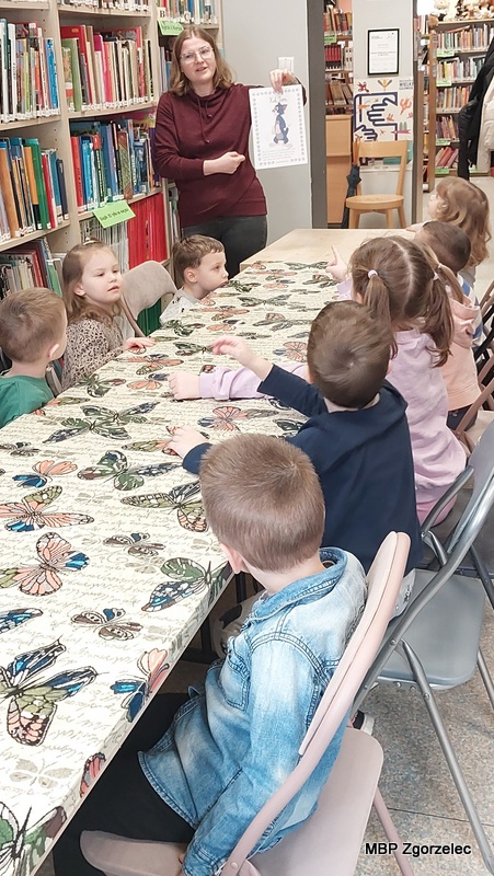 Dzieci siedzą przy stole w&nbsp;czytelni biblioteki, a&nbsp;bibliotekarka stoi obok regału i&nbsp;prezentuje ilustrację z&nbsp;kotem. Dzieci patrzą w&nbsp;stronę pokazywanej ilustracji i&nbsp;siedzą przy stole nakrytym obrusem we wzór motyli. W&nbsp;tle widoczne są regały z&nbsp;książkami oraz wejście do dalszej części biblioteki. Zdjęcie wykonano 23 lutego 2026 r. jako dokumentacja zajęć edukacyjnych.